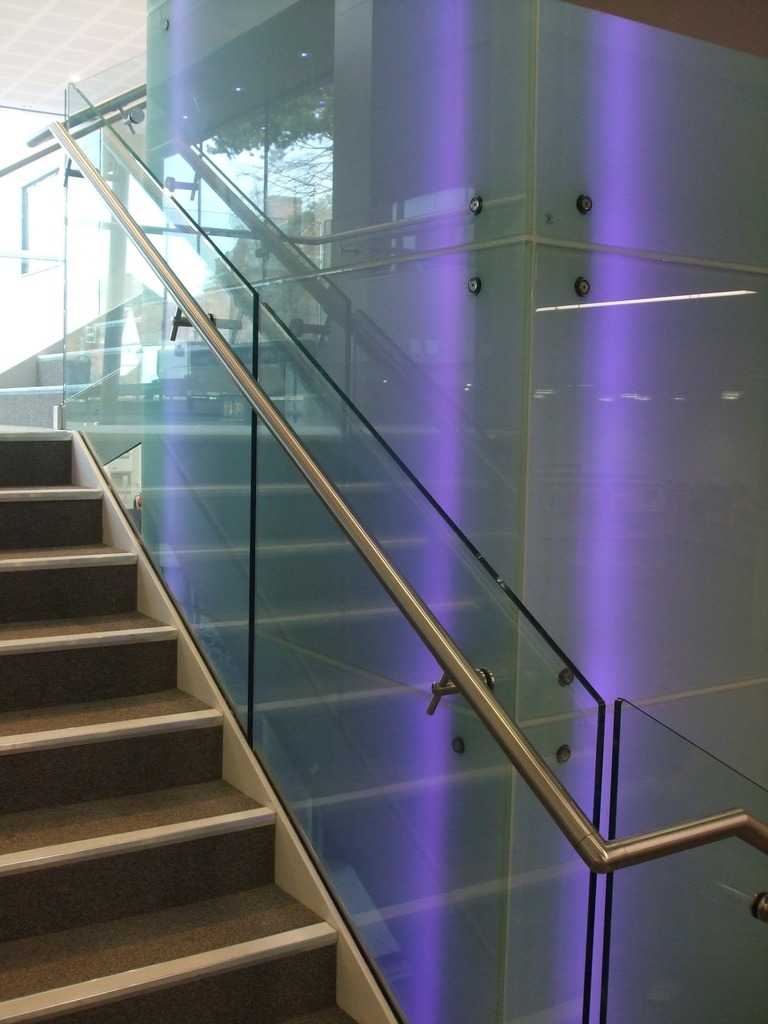 Offset bracket bolted through a glass panel carrying a round tube handrail
