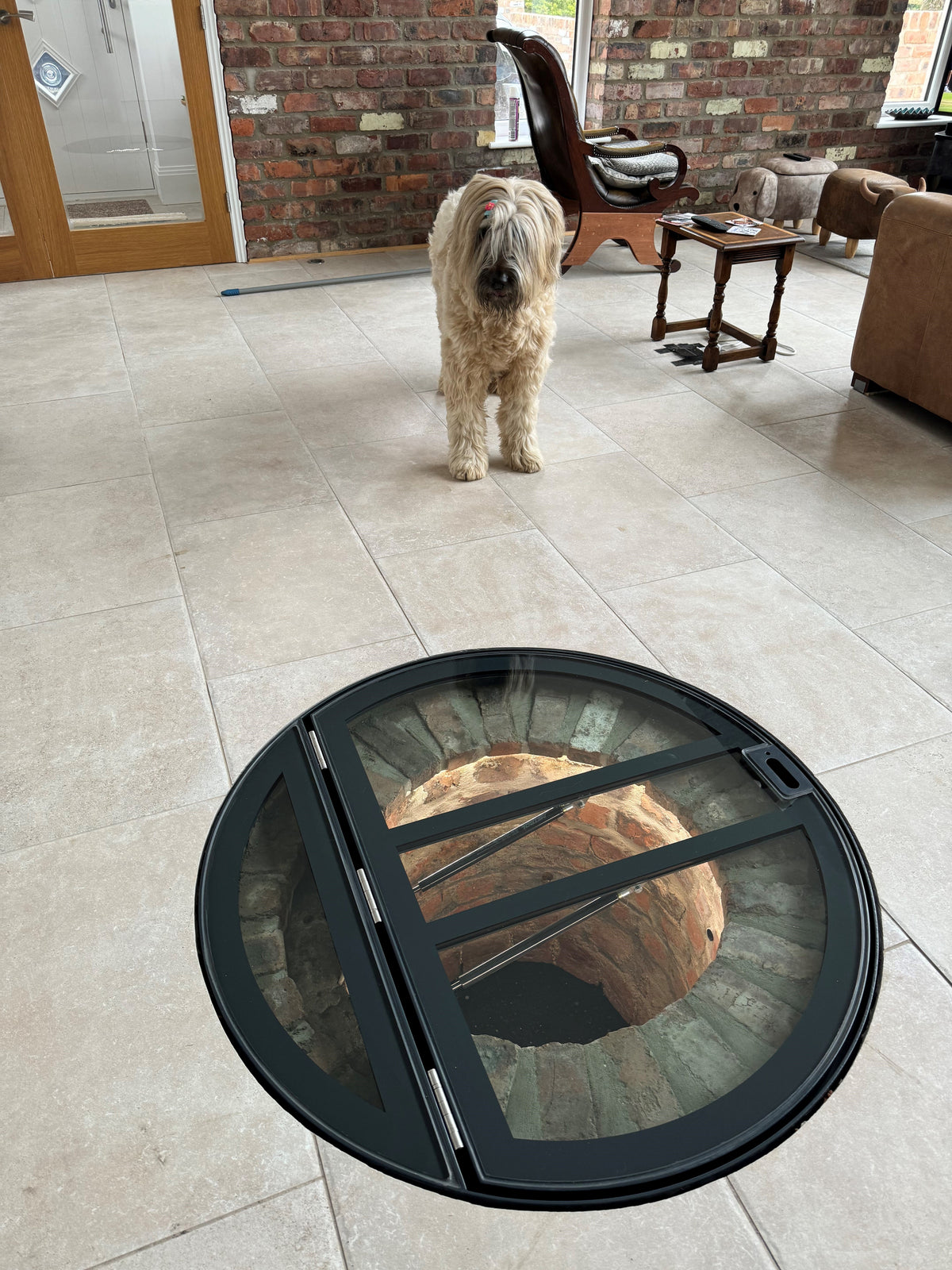 Circular glass wine cellar door set into a tiled floor, the opening leaf carried on twin gas struts with a fixed panel at the rear