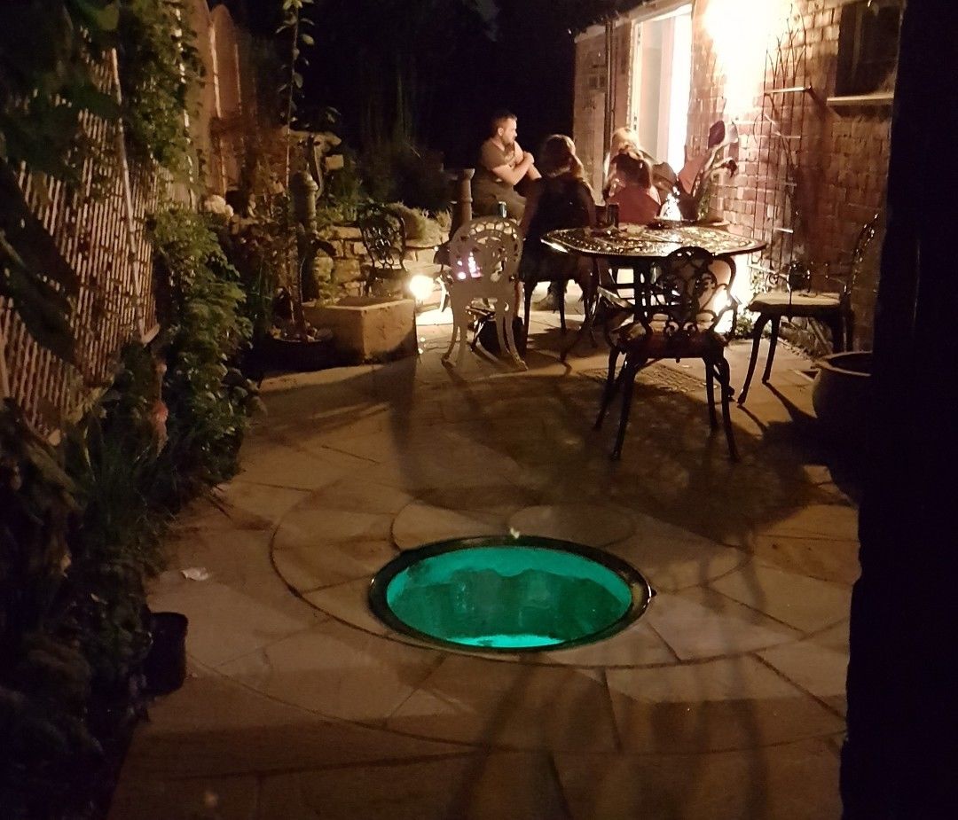 Lit circular glass well cover at night on a patio with garden seating